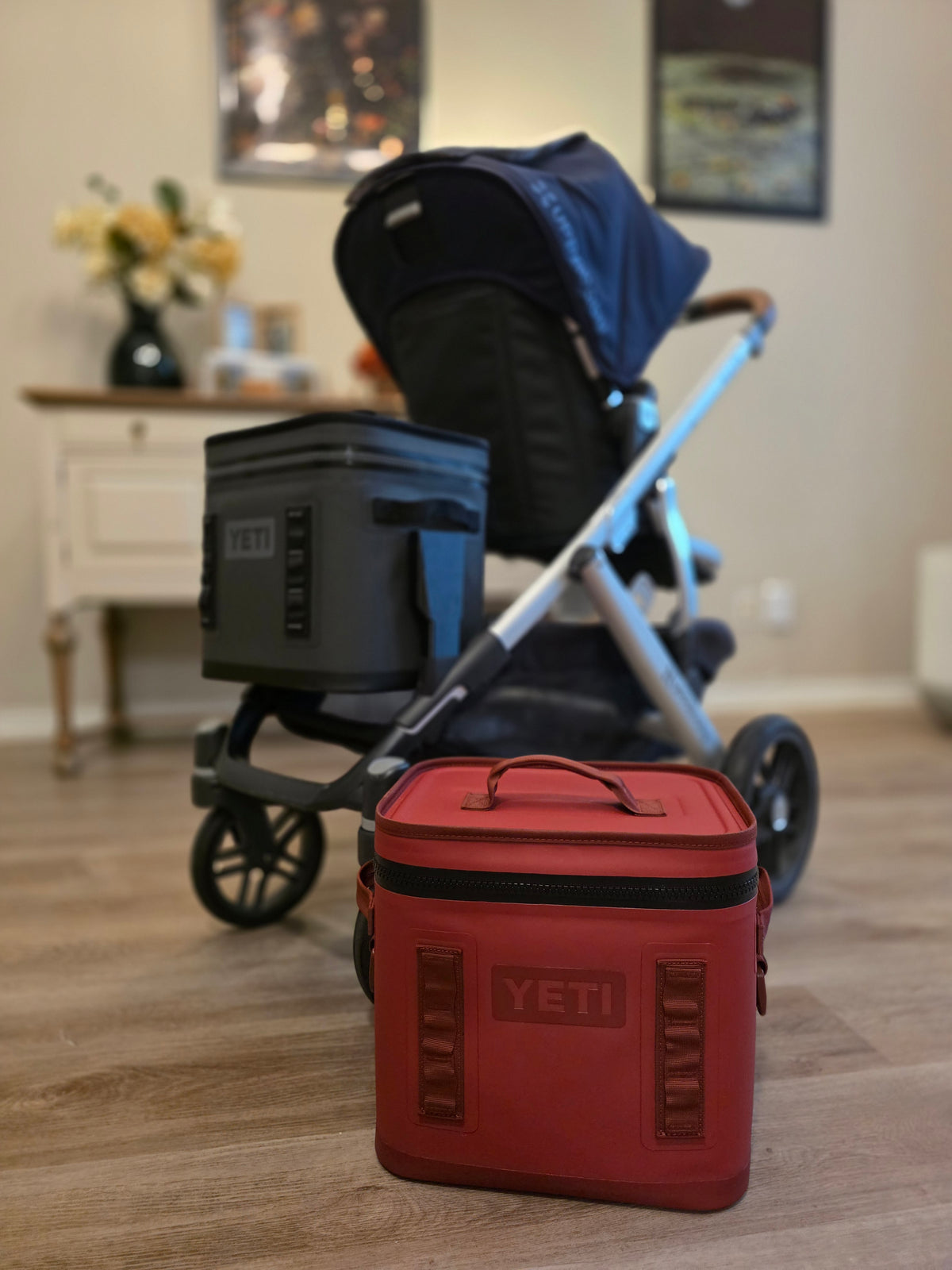 A red YETI Hopper Flip standing on the floor beside an UPPAbaby Vista that is carrying a charcoal one on the cooler mount