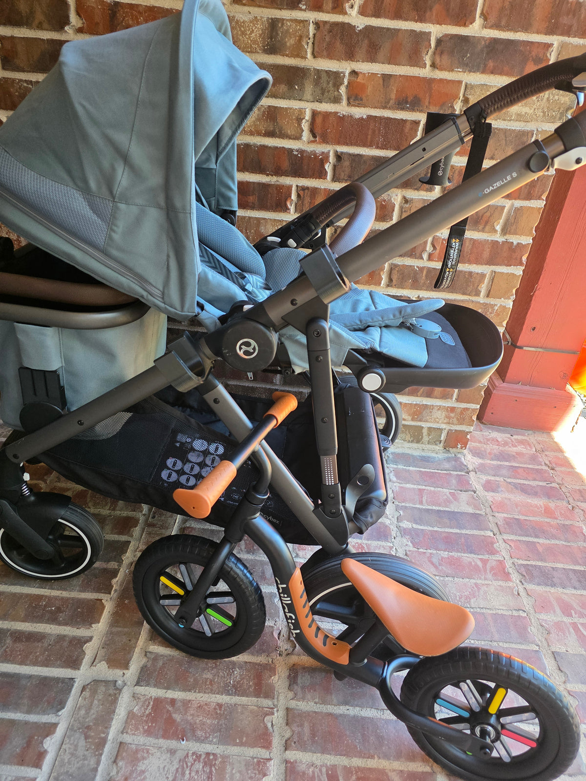 A light blue CYBEX eGazelle S parked against a brick wall with a black and tan balance bike carried on the side of the frame, its wheels hanging clear of the ground.