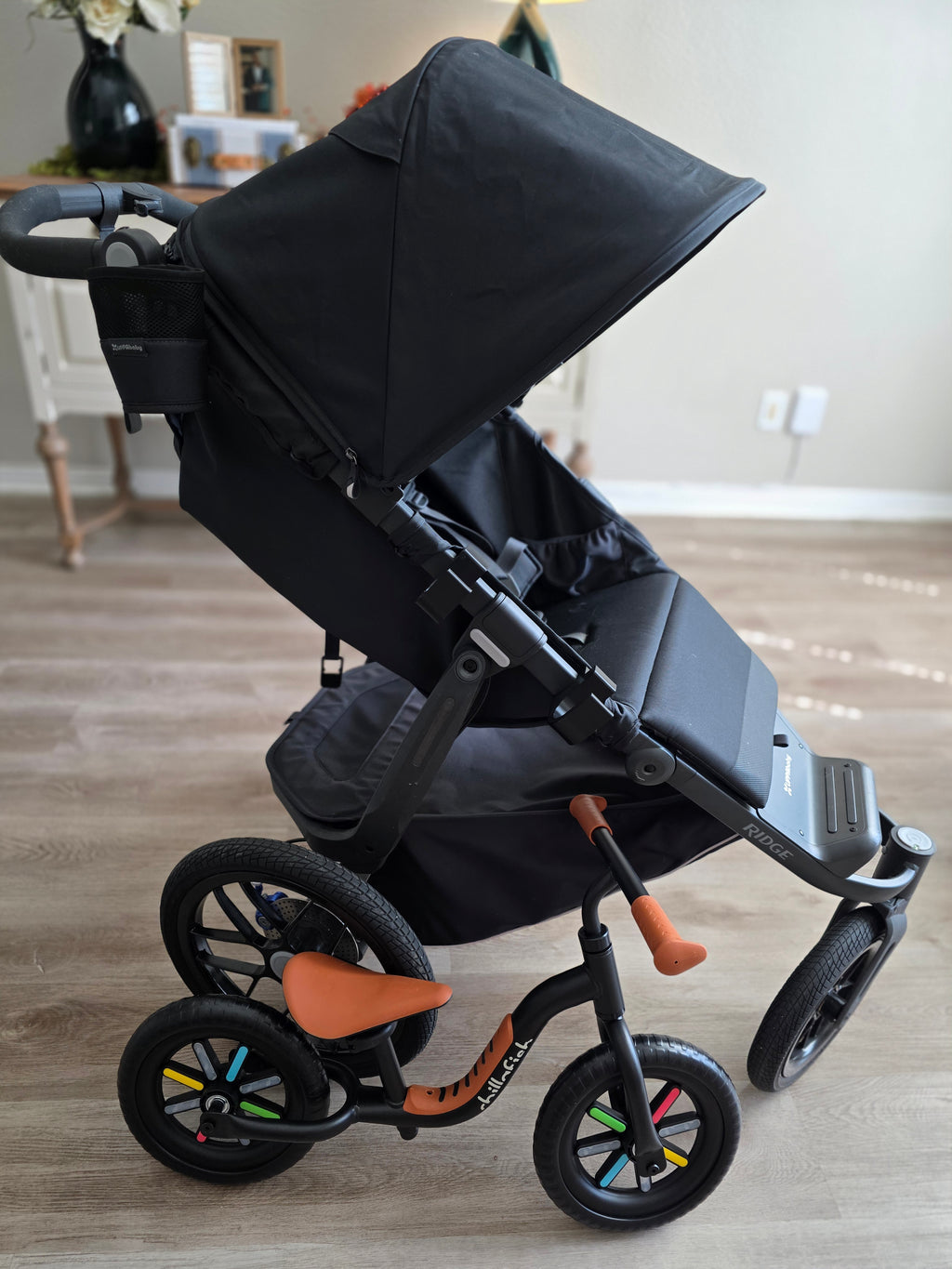 A black UPPAbaby Ridge stroller indoors with a black and tan balance bike standing beside it.
