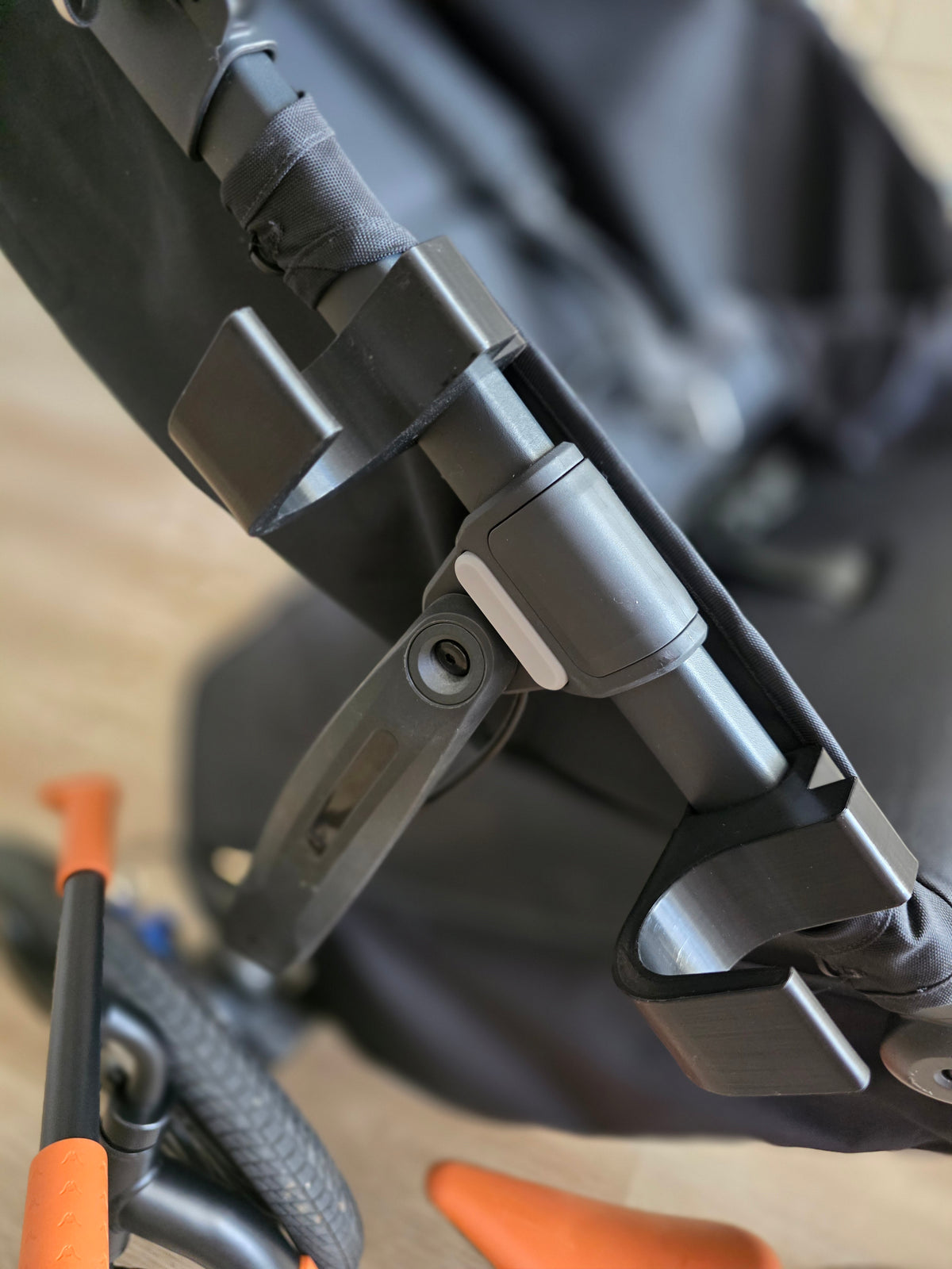 Detail of the black Stroller House holders hooked over the UPPAbaby Ridge frame tube, showing the deep top hook and the open U-shaped cradle.