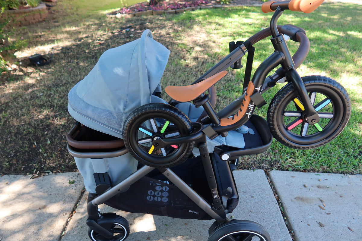 A CYBEX eGazelle S on a sidewalk with a black and tan balance bike riding level on the side of the frame, held by a pair of black Stroller House holders.
