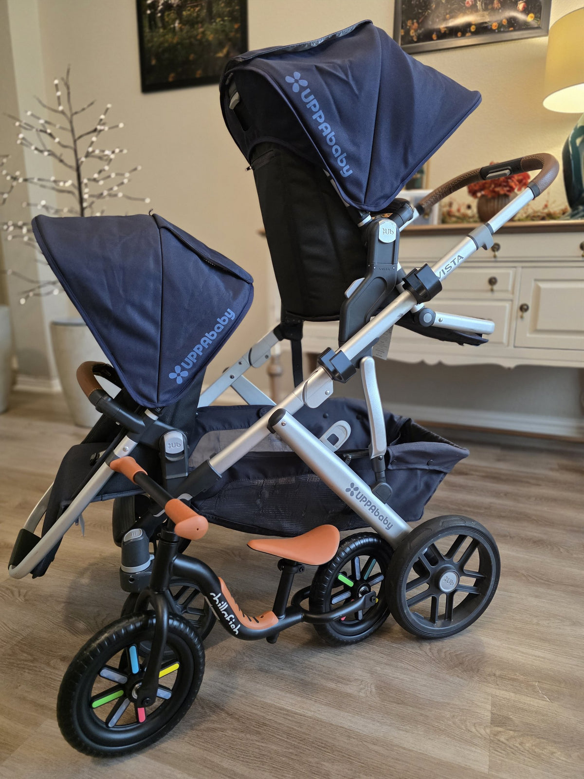 A navy UPPAbaby Vista with a black and orange balance bike carried on the left side of the frame by a pair of black Stroller House holders, hanging clear of the wheels.